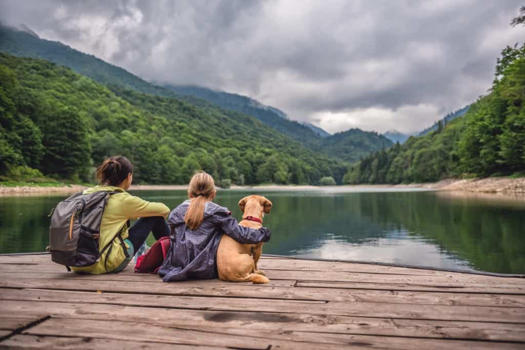 Mother and daughter with a dog resting on a pier looking at lake