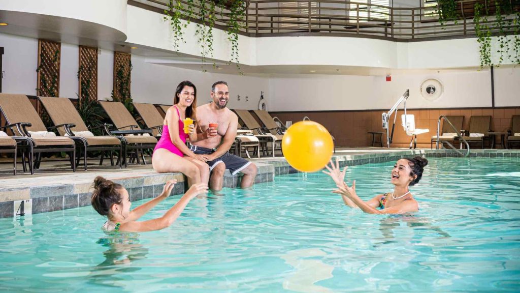 Couple sits on edge of pool with 2 kids playing in water.