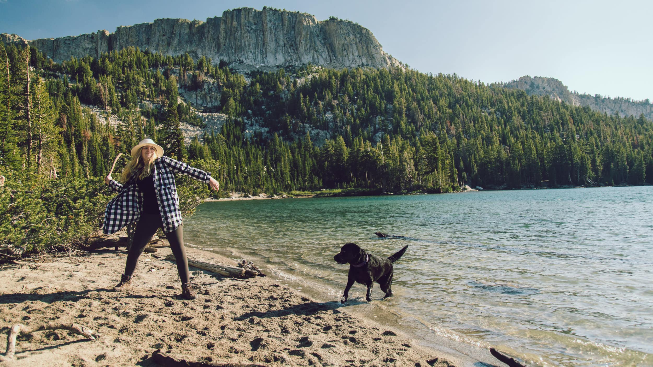 woman tosses stick to black lab at edge of alpine lake