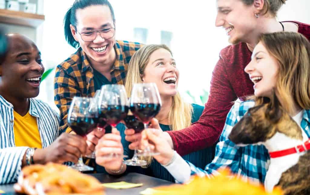 group of friends sit at a table with a dog clinking full wine glasses