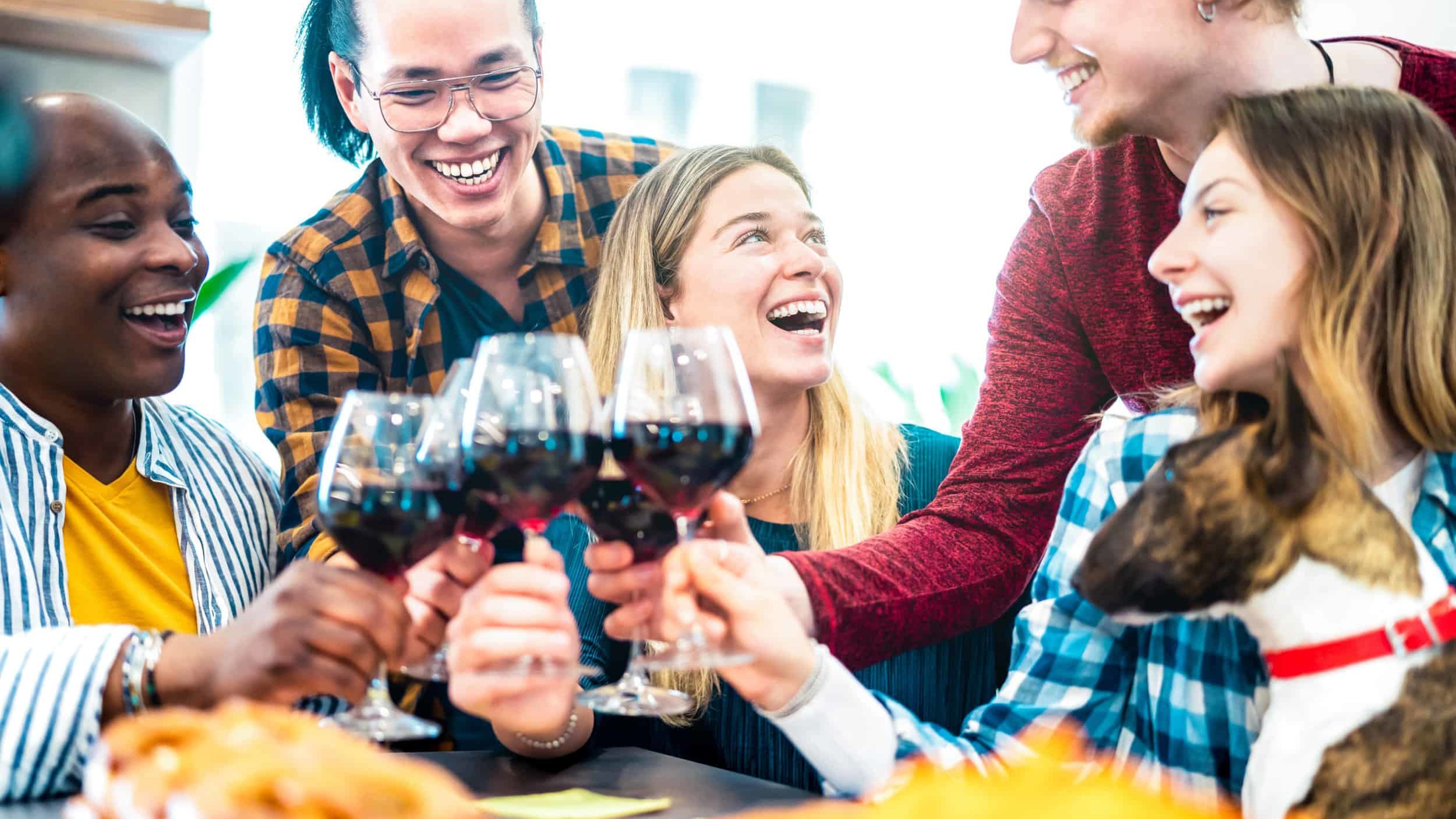 group of friends sit at a table with a dog clinking full wine glasses