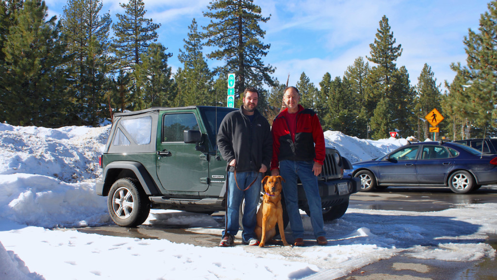 2 men and a dog stand in snowy parking lot